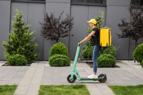 Volledige lengte schot van jonge levering vrouw in gele cap met thermo zak of rugzak rijden een scooter — Stockfoto