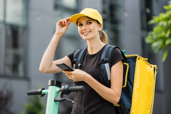 Attractive female courier in yellow cap look at the camera, searching customer on smartphone. — Fotografia de Stock