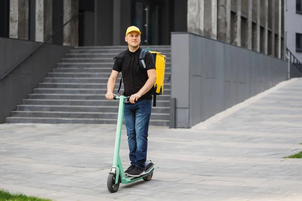 Happy delivery man met gele pet en thermische geïsoleerde tas paardrijden elektrische scooter op straat. — Stockfoto