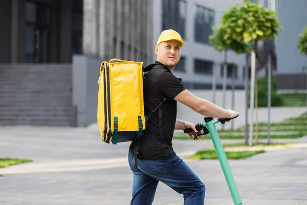 Happy delivery man σε κίτρινο καπάκι και θερμική μόνωση τσάντα ιππασίας ηλεκτρικά σκούτερ στην οδό της πόλης. — Φωτογραφία Αρχείου