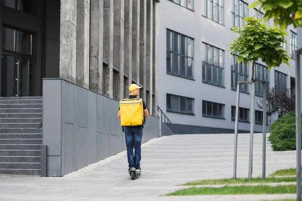 Sırtında sarı bir sırt çantası ve günlük yaz kıyafetleri olan genç bir adam elektrikli scooterla hareket ediyor.