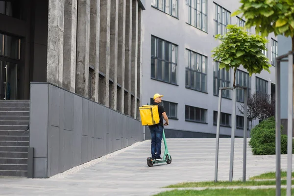 Sırtında sarı bir sırt çantası ve günlük yaz kıyafetleri olan genç bir adam elektrikli scooterla hareket ediyor.