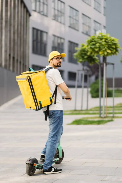Happy delivery man in yellow cap and thermal insulated bag riding electric scooter on city street. — Fotografia de Stock