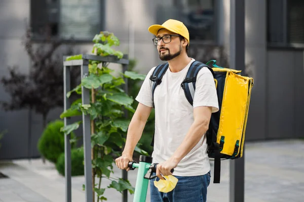 Rijder man in gele pet en thermische doos rugzak leveren maaltijd aan klanten met elektrische scooter. — Stockfoto