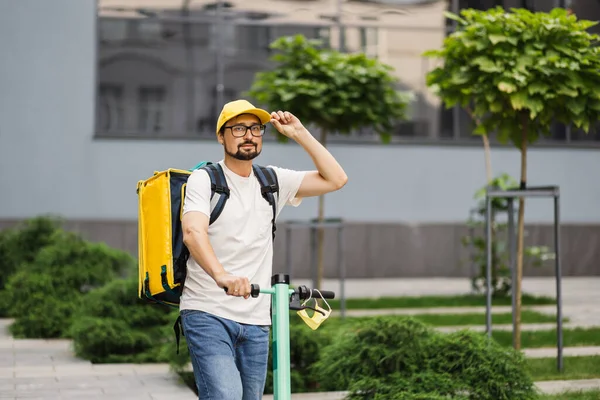 Beyaz erkek kurye scooterla şehirde sıcak yiyeceklerle yemek dağıtıyor.