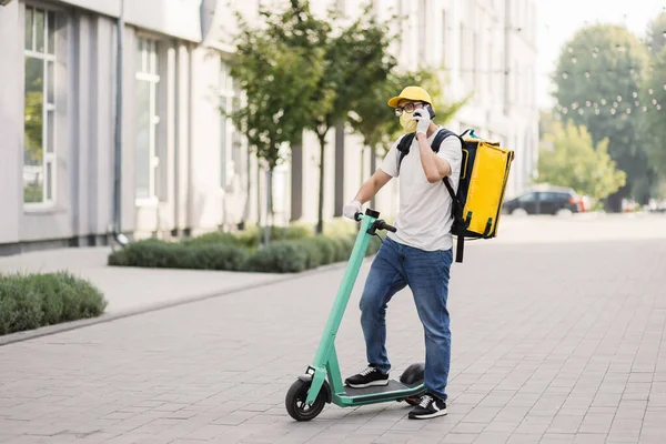 Akıllı telefon kullanan ve şehirde scooter kullanan yüz maskeli bir teslimatçının yan görüntüsü.