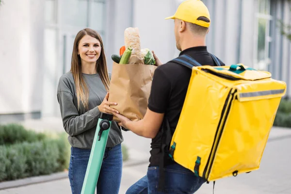 Young man on scooter with thermal bag gives paper bag with fresh vegetables to woman client.