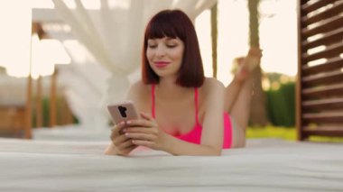 Odaklanmış, pembe mayo giymiş beyaz bir kadın tatil köyü havuzunun yanındaki ahşap bir çardakta yatıyor.