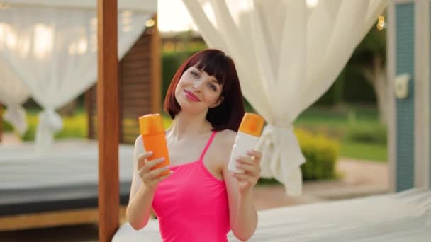 Жінка, що сидить на пляжному ліжку біля басейну пропонує вибрати трубки з сонцезахисним кремом або соляним лосьйоном — стокове відео