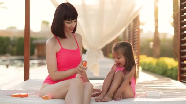 Молода красива жінка наносить сонцезахисний спрей на ноги дочки в рожевих купальниках — стокове відео
