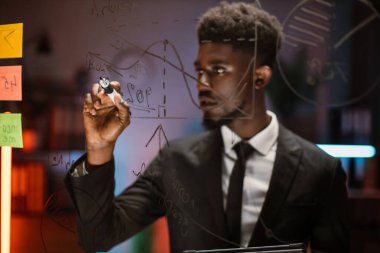 Afro-Amerikalı adam, cam tahtada yazıyor ve proje planı analizi yapıyor.