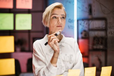 Yeni proje stratejisi üzerinde çalışan iş kadını, yapışkan notlarla cam tahtanın yanında duruyor.