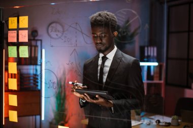 Erkek Afro-Amerikan finans uzmanı elinde tablet tutarak ofiste cam duvarın yanında duruyor.