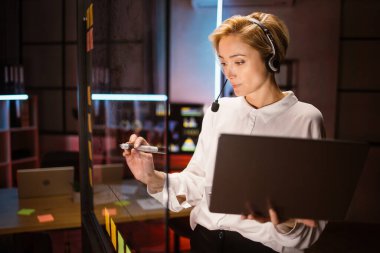 Kadın işçi, kulaklık takıyor, dizüstü bilgisayar kullanıyor, fikirler yazıyor ve cam duvara projeler yazıyor.