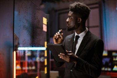 Cam tahtanın önünde elinde tablet bilgisayarla duran Afro-Amerikan erkek girişimci.