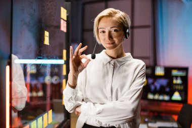 Beyaz kulaklı kadın, keçeli kalemle şeffaf cam tahtanın yanında poz veriyor.