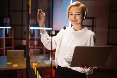 Kadın işçi, kulaklık takıyor, dizüstü bilgisayar kullanıyor, fikirler yazıyor ve cam duvara projeler yazıyor.