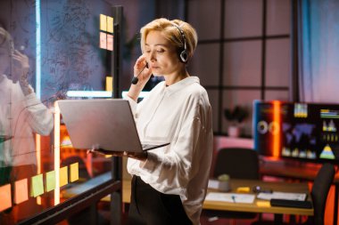 Kafkasyalı iş kadını, video görüşmesi için dizüstü bilgisayar kullanıyor, cam tahtanın yanında duruyor.