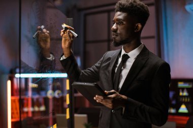 Afrika kökenli Amerikalı finans uzmanı, çalışma planını kontrol ederken tablet kullanıyor, cam levhaya bakıyor.