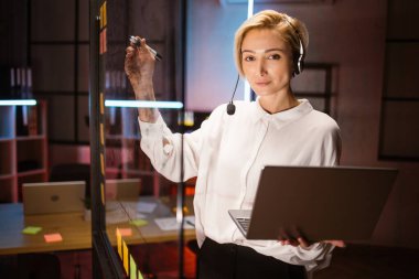 Kadın işçi, kulaklık takıyor, dizüstü bilgisayar kullanıyor, fikirler yazıyor ve cam duvara projeler yazıyor.