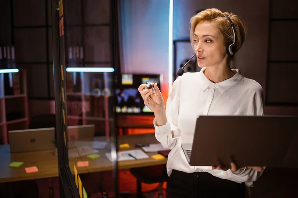 Female manager, wearing headset and using laptop, standing near glass wall in modern dark office —  Fotos de Stock