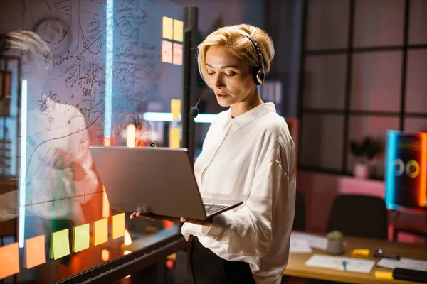 Kafkasyalı iş kadını, video görüşmesi için dizüstü bilgisayar kullanıyor, cam tahtanın yanında duruyor.