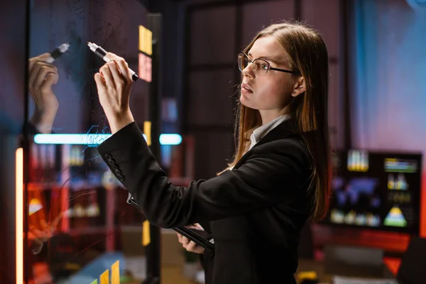 Businesswoman in eyeglasses drawing on glass board with stickers, working on project tasks — Fotografia de Stock