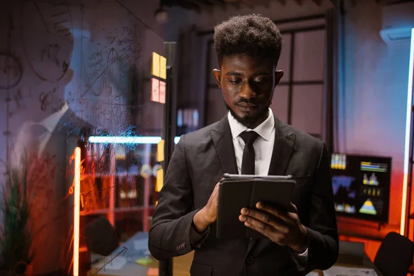 ชายชาวแอฟริกันอเมริกันจัดการโครงการบนแท็บเล็ตพีซี ทํางานในออฟฟิศกลางคืนกับกระดานกระจกสกัม ภาพถ่ายสต็อกที่ปลอดค่าลิขสิทธิ์