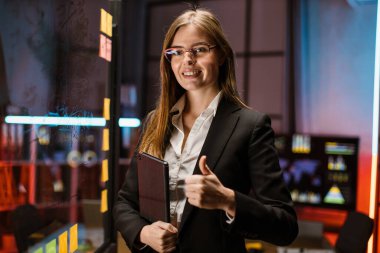 Kadın yönetici, elinde tablet bilgisayar, yapışkan kağıtlarla cam duvarın yanında duruyor.