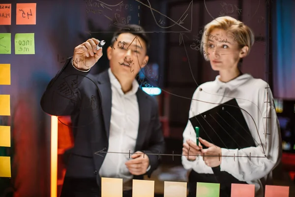 Asian businessman brainstorming, talking with female colleague, standing behind glass board — Fotografia de Stock