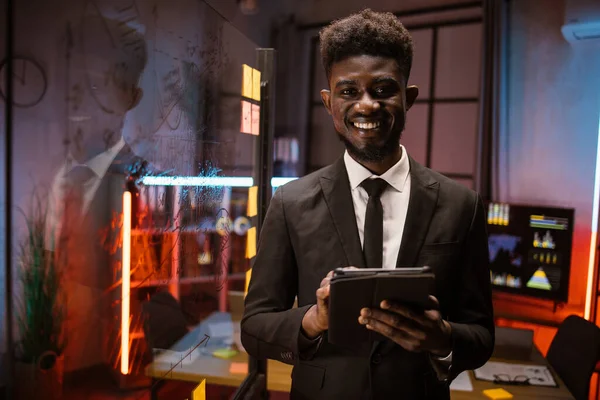アフリカ系アメリカ人男性タブレットPC上でプロジェクトを管理し、スクラムガラスボードで夜間オフィスで働く ストック画像