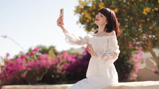 Mujer con sombrero y vestido blanco usando un teléfono inteligente o tomando una selfie en la ciudad tropical en verano — Vídeos de Stock