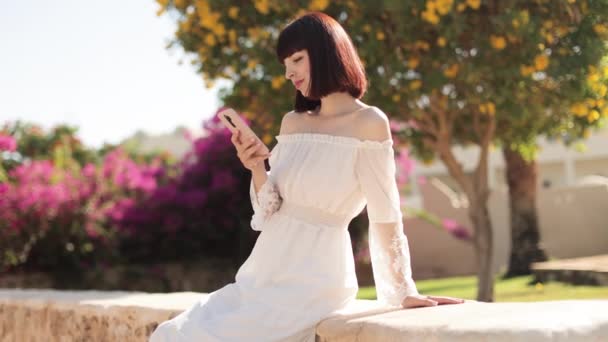 Mujer con sombrero y vestido blanco usando un teléfono inteligente o tomando una selfie en la ciudad tropical en verano — Vídeos de Stock