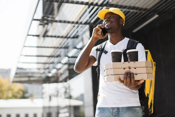 Young African American courier, pizza and coffee delivery man with thermal backpack and phone. — Stockfoto