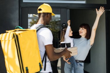Erkek Afrikalı kurye pizza kutuları veriyor ve kahveyi restorandan alıp gülümseyen mutlu aileye götürüyor.