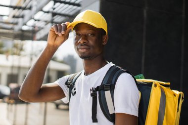 Portrait of delivery man with yellow cap and thermal backpack looking at camera.