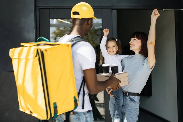 Erkek Afrikalı kurye pizza kutuları veriyor ve kahveyi restorandan alıp gülümseyen mutlu aileye götürüyor.