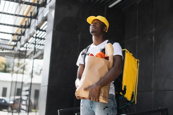 Voedsellevering Afrikaanse man met papieren zak met biologische verse groenten met gele dop en rugzak — Stockfoto