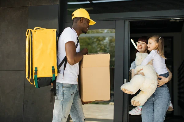 Young caucasian woman with her little girl receiving a package with teddy bear from African courier — Foto de Stock