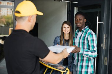Sarı üniformalı teslimatçı. Paket kutusunu alıcıya teslim ediyor. Genç kadın ve kocası.