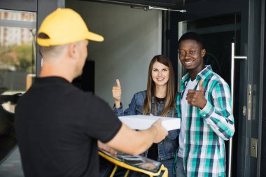 Sarı üniformalı teslimatçı. Paket kutusunu alıcıya teslim ediyor. Genç kadın ve kocası.