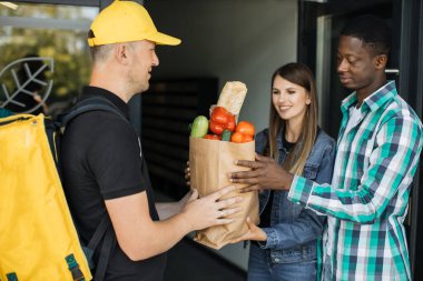 Sarı üniformalı beyaz teslimatçı. İçinde market yemekleri olan kağıt eko çanta getiriyor.