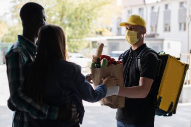 Happy food delivery man with thermal backpack delivers paper bag with fresh vegetables order