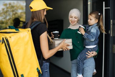 Kadın kurye, gülümseyen Müslüman aileye pizza kutuları ve kahve veriyor.