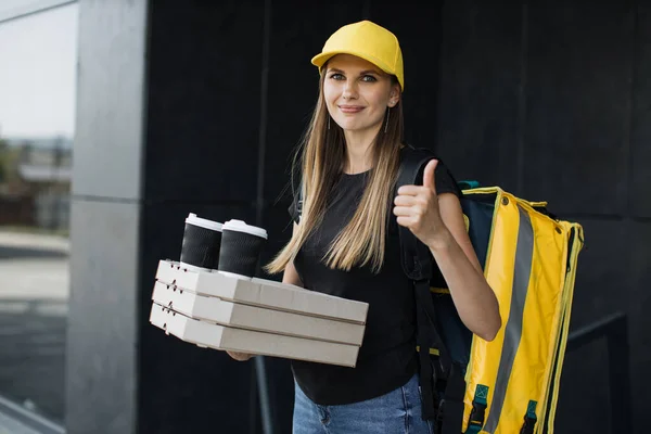 Delivery woman in yellow cap and thermo bag or backpack waiting for the customer near building