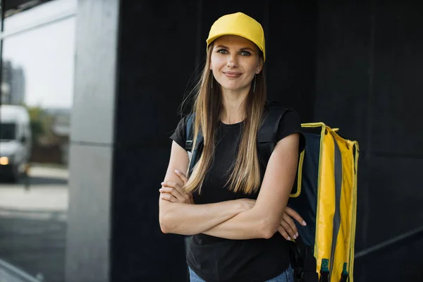 Delivery woman in yellow cap and thermo bag or backpack waiting for the customer near building