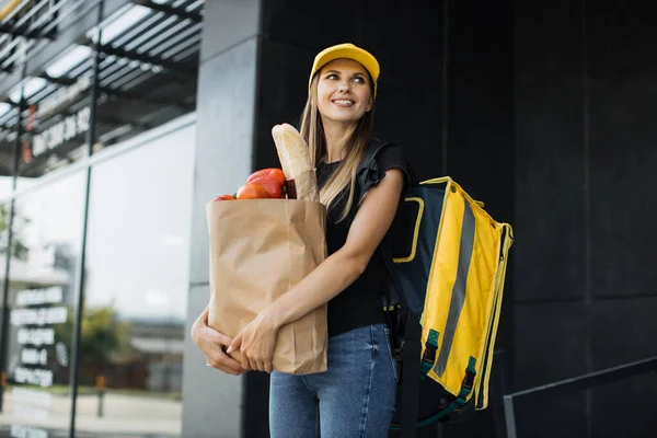 Entrega de alimentos mulher segurando saco de papel com vegetais frescos orgânicos com tampa amarela e mochila — Fotografia de Stock