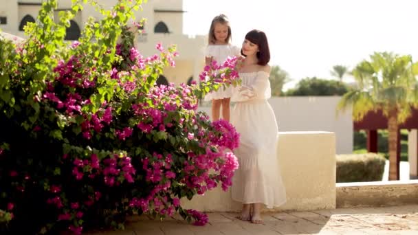 Smiling mom with daughter on white dresses sitting against the background of a tropical houses — Vídeos de Stock