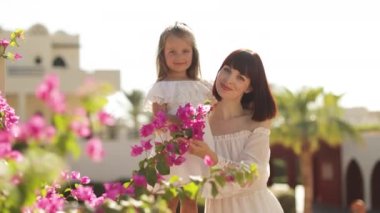 Beyaz elbiseli bir kızla gülümseyen anne, tropik evlerin arka planında oturuyor.