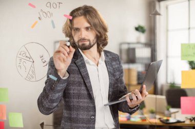 Erkek çalışan dizüstü bilgisayar kullanıyor ve cam panoya yazı yazıyor.